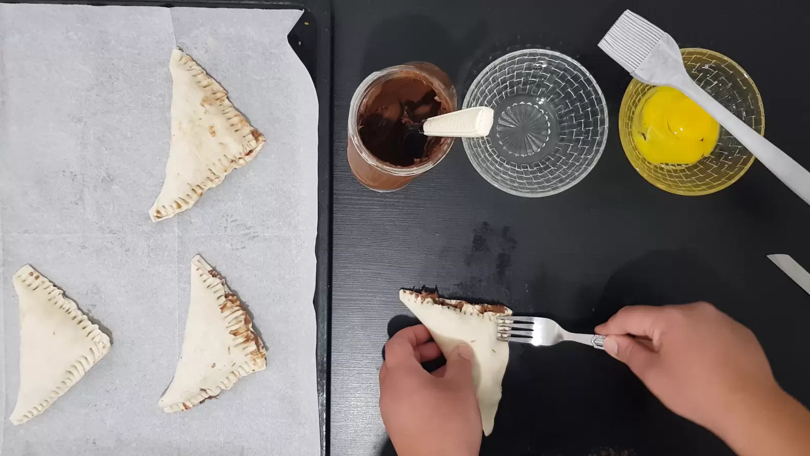 A bowl with an egg and a brush for spreading, an empty bowl, chocolate spread, a baking sheet with baking paper and puff pastry triangles on it and a fork