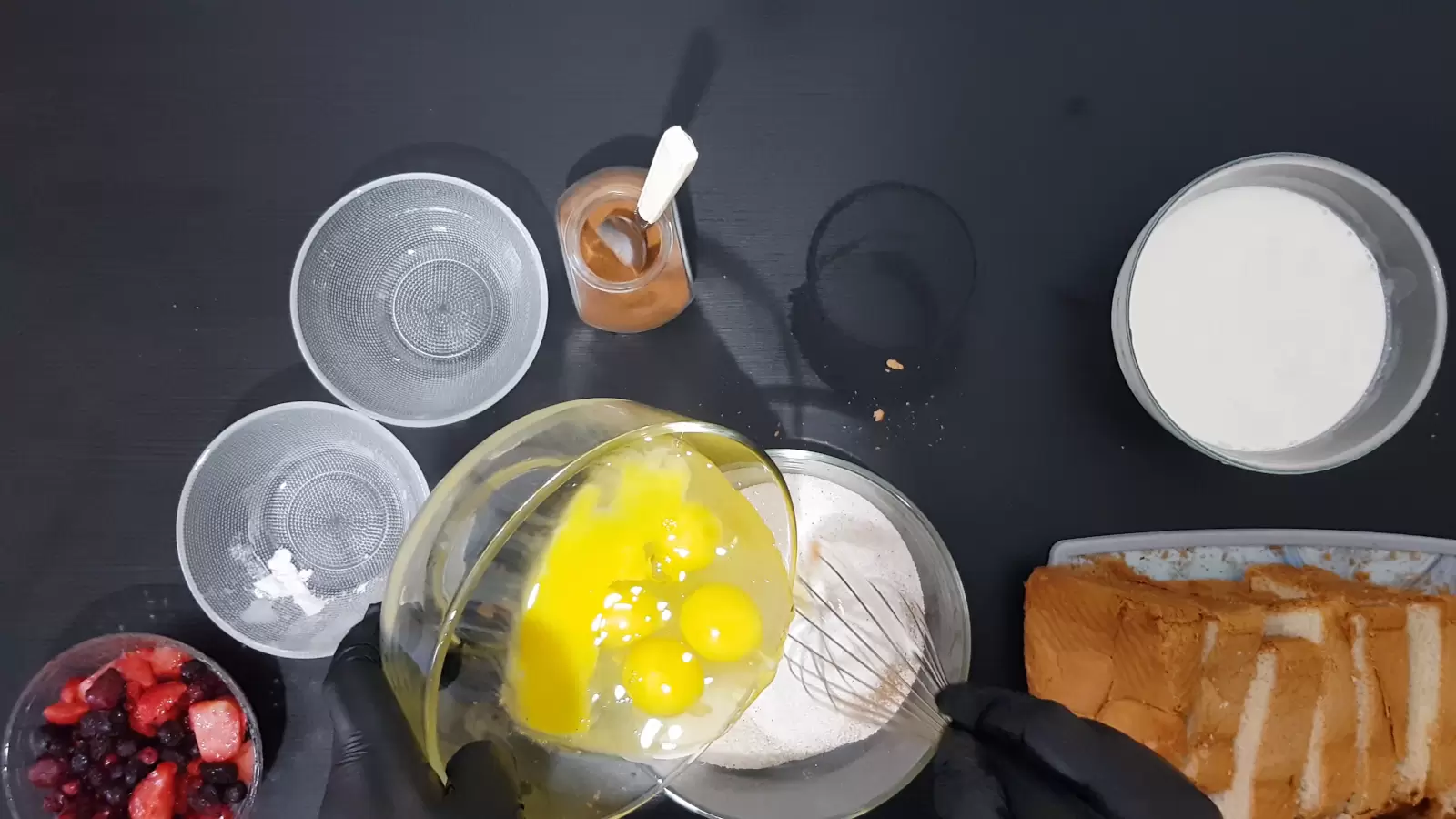 Bowl of heavy cream, bowl of eggs spilling into bowl With batter, jar with cinnamon spice and spoon, 2 empty bowls, bowl with berries, sliced ​​bread on cutting board, whisk