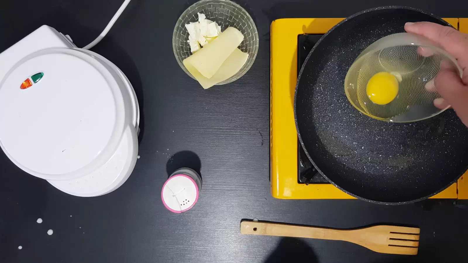 A bowl with an egg spilled into the pan, a bowl with cheeses. Waffle maker closed.