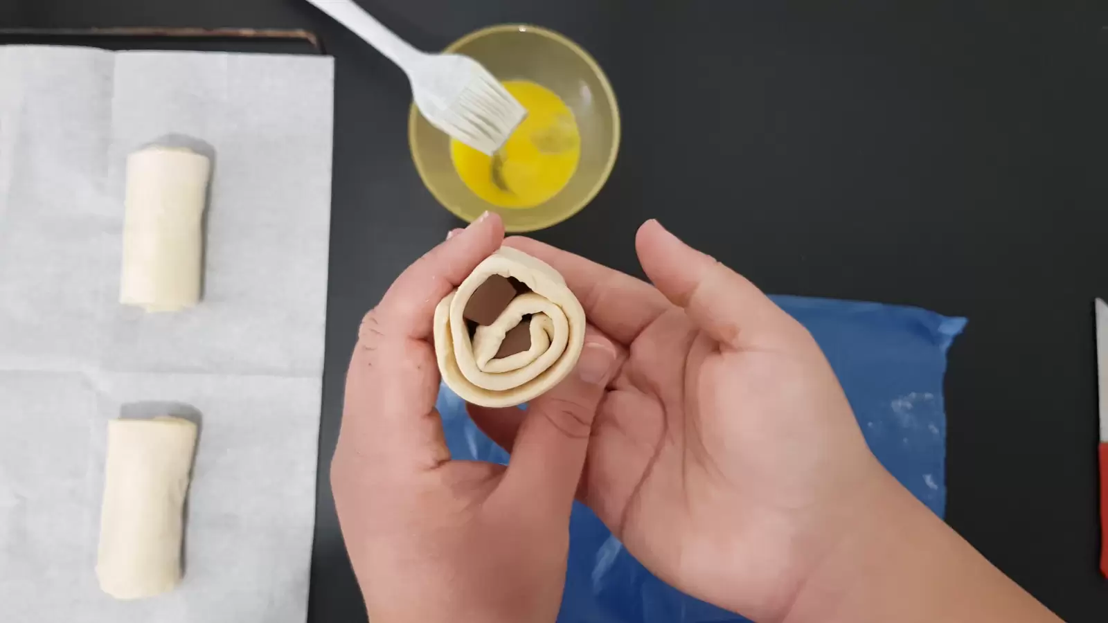 Puff pastry rolled with Kinder Bueno, a bowl with an egg and a brush, baking paper in an oven tray