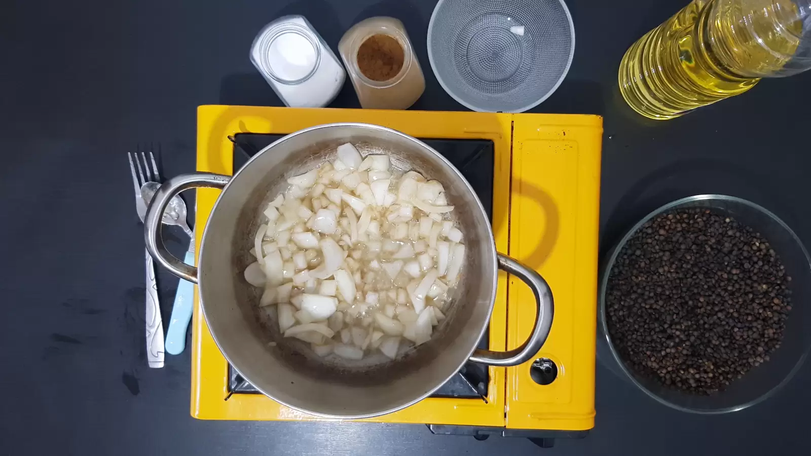 Bowl with lentils, empty bowl, jar of cumin, jar of salt, bottle of oil, gas jar, pot with chopped onion, knife and fork