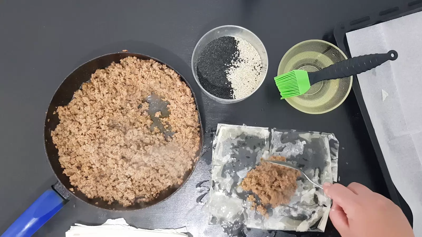 bowl with oil and brush, bowl with white and black sesame, pan with beef, sheet of phyllo with beef