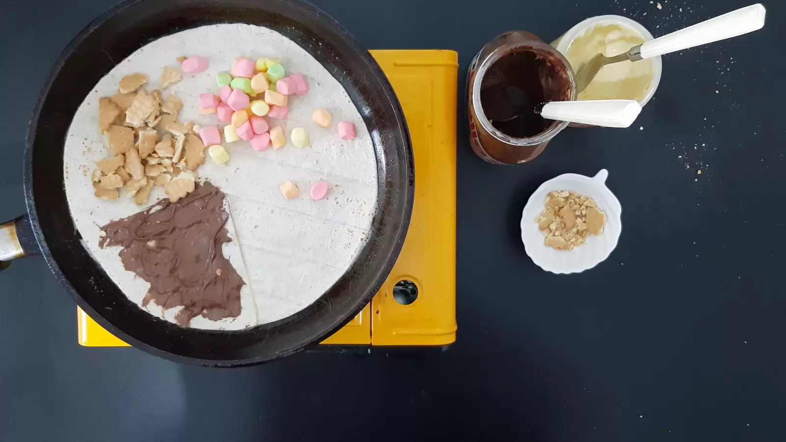 A bowl of biscuits, a jar of white chocolate, a jar of Nutella chocolate, a tortilla in a pan with chocolate, biscuits and marshmallows