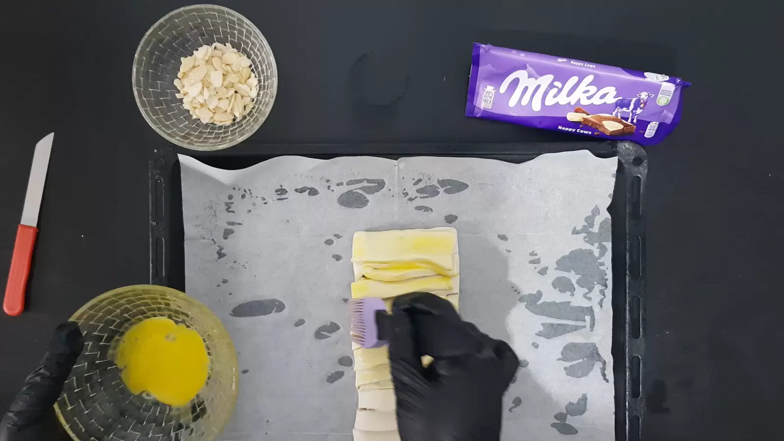 pack of Milka chocolate, a brush for spreading in a bowl with egg, a bowl with almonds, puff pastry with Milka chocolate on a baking sheet with baking paper, a knife