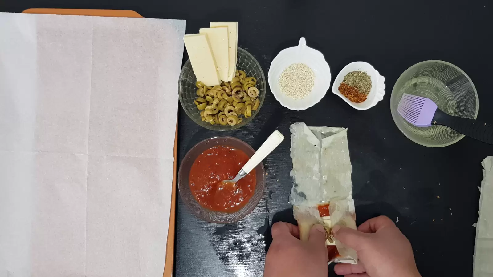 Bowl with oil and brush, bowl with spices, bowl with sesame seeds, bowl with olives and yellow cheese, bowl with tomato sauce, filo dough, spoon and baking sheet with baking paper