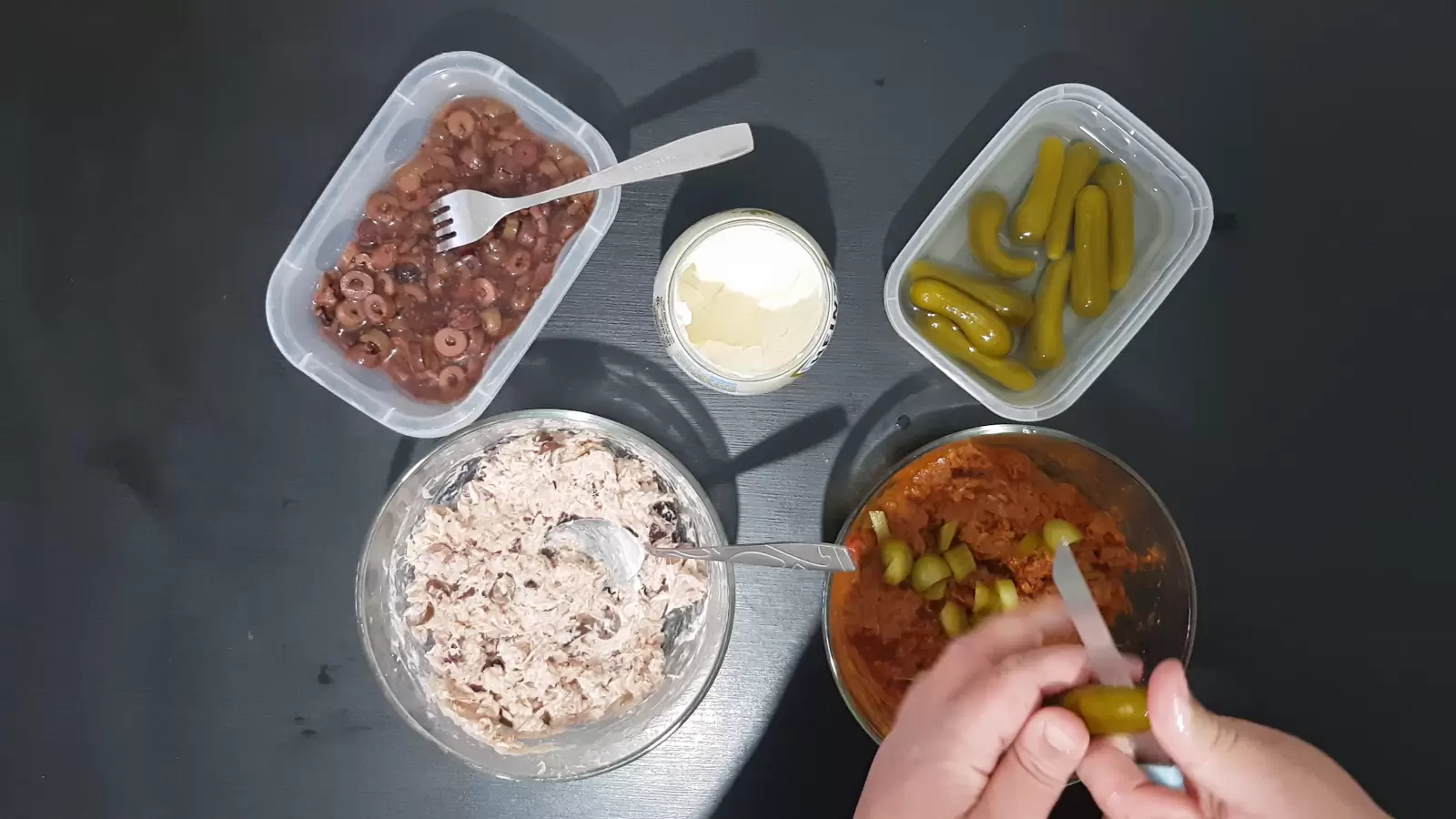 A box with pickles, a box with olives, a spoon with mayonnaise, a fork, a knife that cuts pickles, a bowl with tuna, a bowl with tuna in tomato sauce