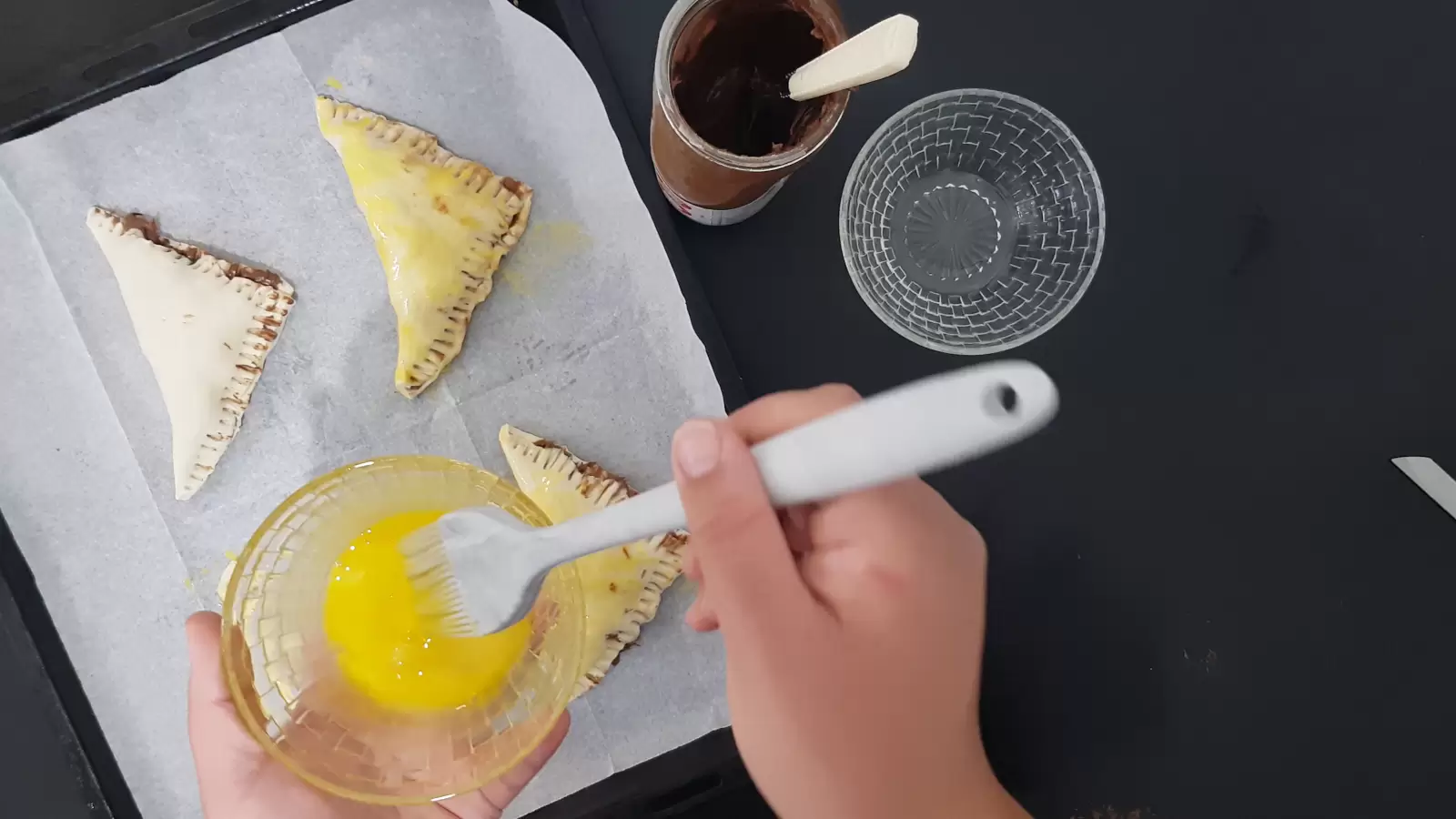 An empty bowl, chocolate spread, a mold with puff pastry, a bowl with an egg and a brush for spreading