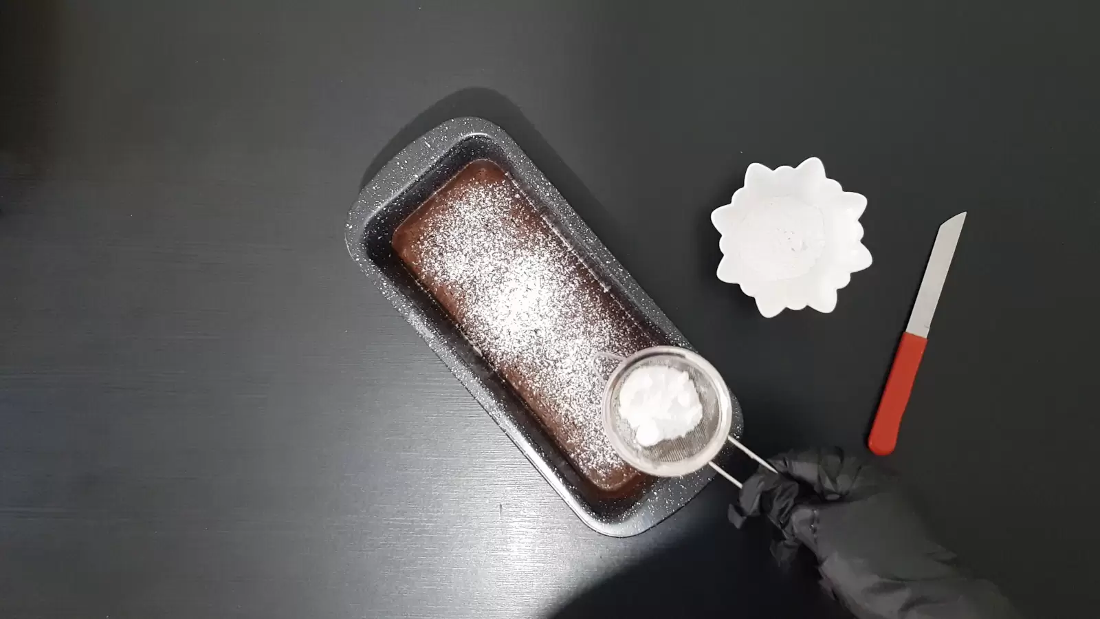 Brownies in pan with powdered sugar on top, bowl with powdered sugar, knife