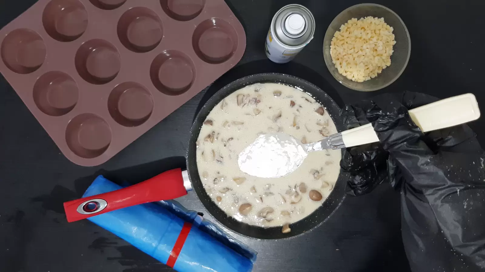 Bowl with grated cheese, oil spray, spoon of flour over pan with mushrooms and cream, muffin tin, rolled filo dough
