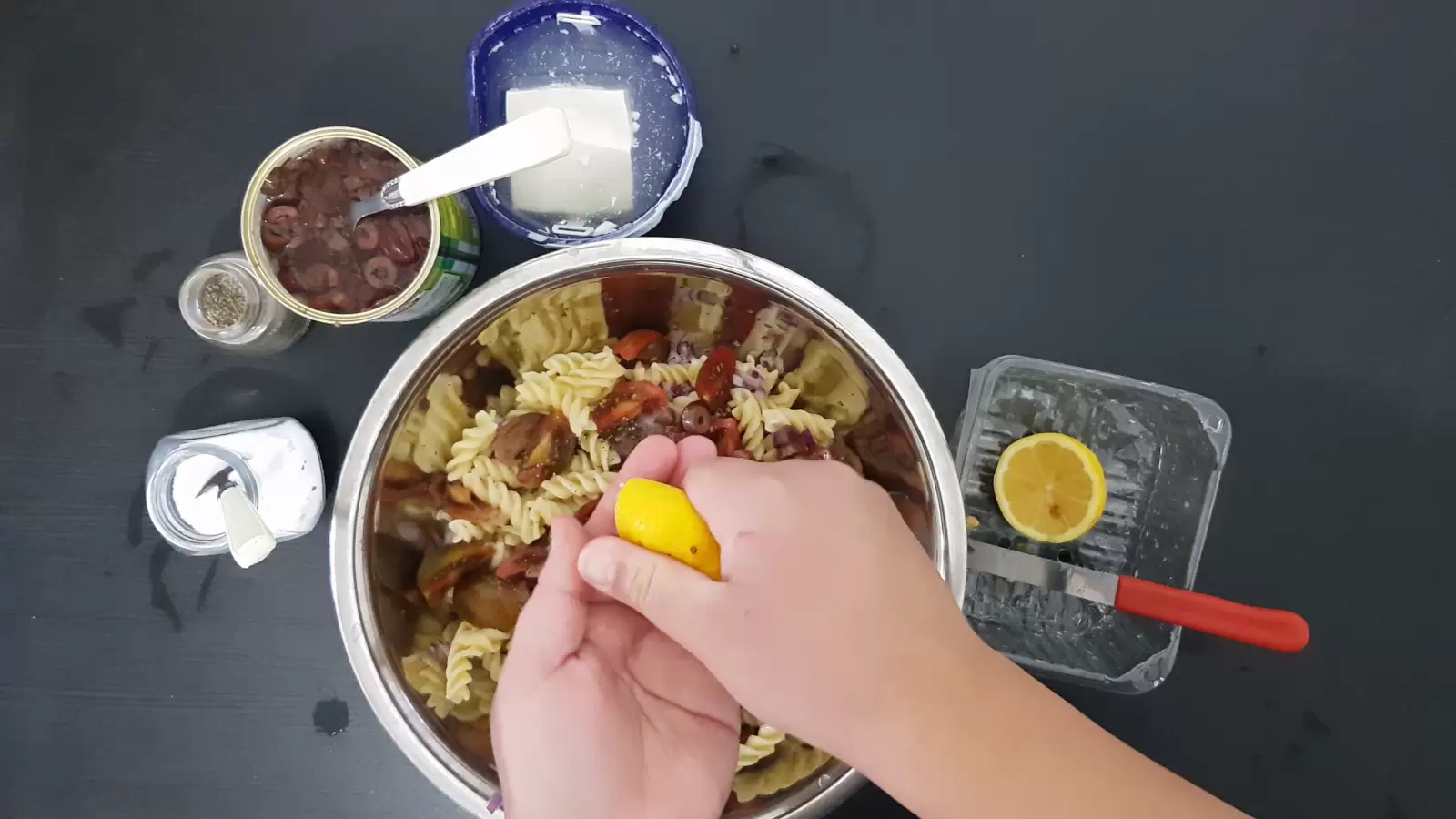 A bowl with pasta and olives, a box with Bulgarian cheese, a can with olives and a spoon, a jar of salt, a jar of pepper and a spoon, a box with a lemon and a knife