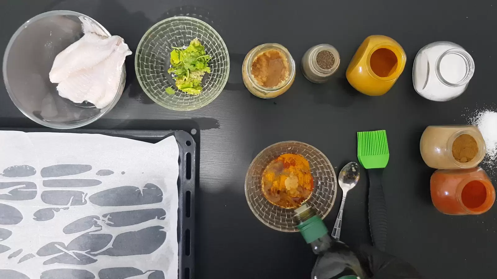 Turmeric, paprika, cumin, pepper, salt, coriander, oven tray, tilapia fish, brush, spoon and olive oil poured into a bowl