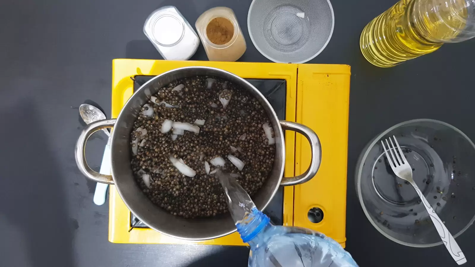 Large empty bowl with fork, small empty bowl, jar of cumin, jar of salt, bottle of oil, gas stove, water pouring from bottle into pot with lentils and chopped onion, knife and fork