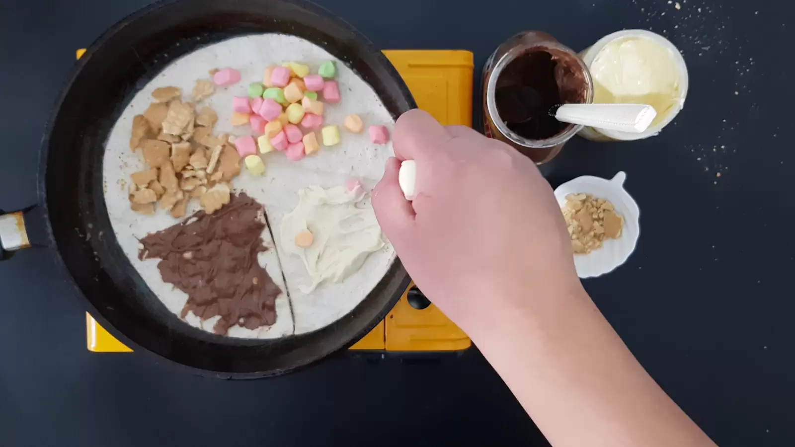 A bowl of biscuits, a jar of white chocolate, a jar of Nutella chocolate, a tortilla in a pan with chocolate, biscuits and marshmallows, a teaspoon of white chocolate spread