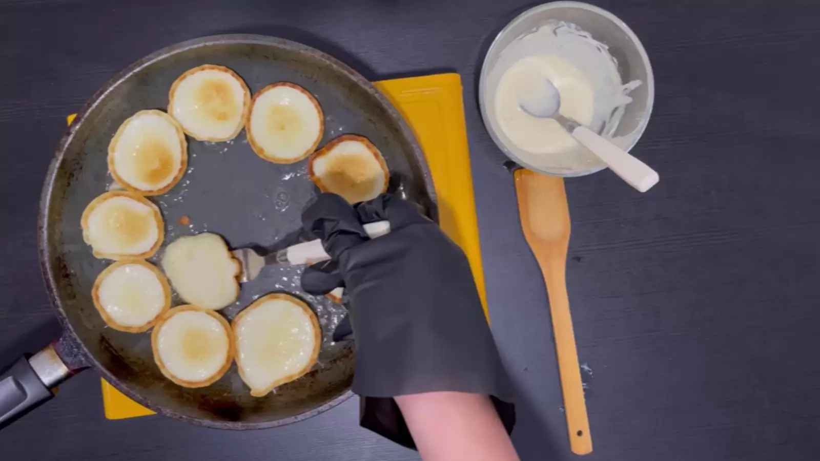 Bowl with batter and spoon, wooden spoon, pan with cheese pancakes and fork