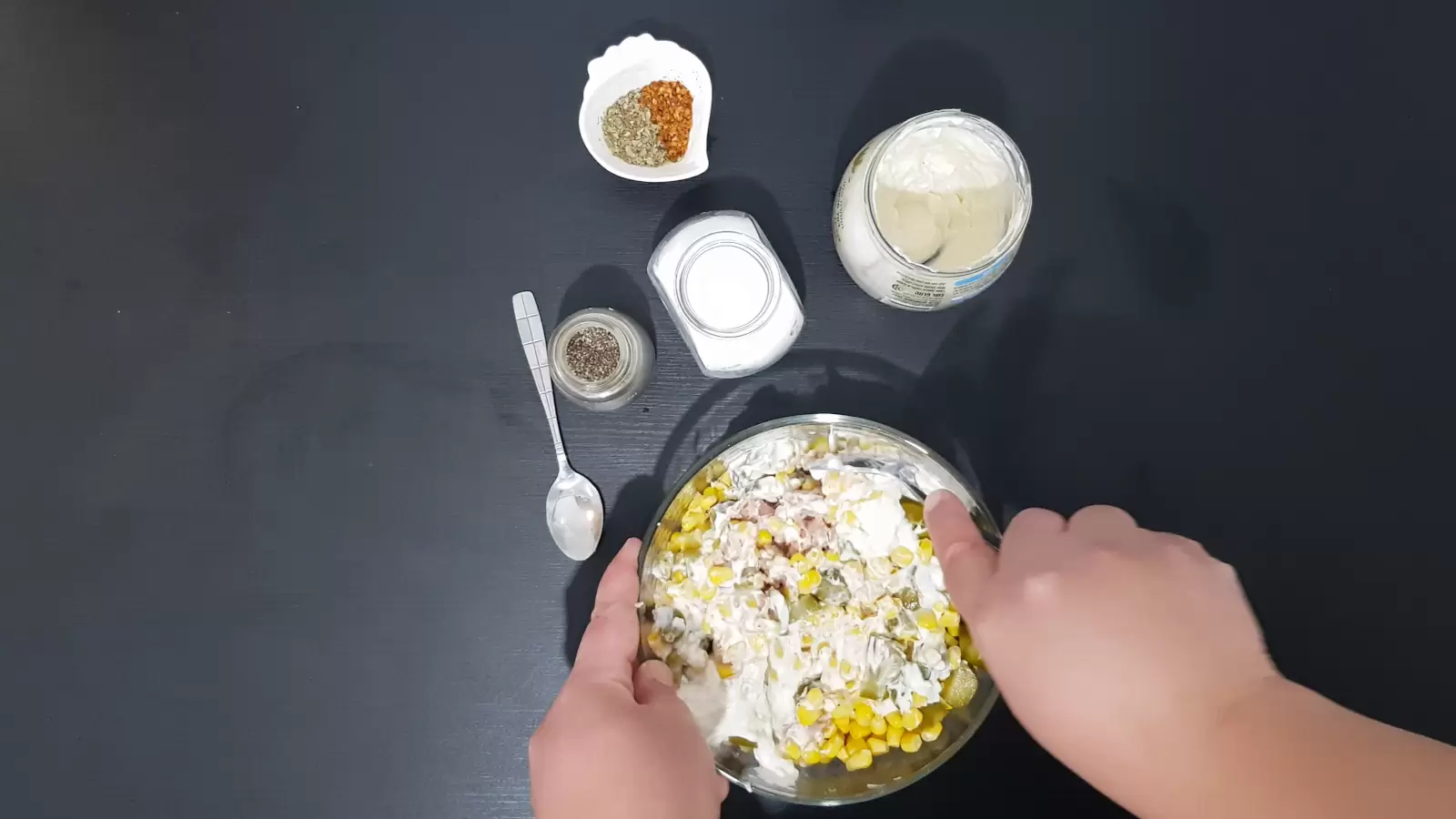 Jar with mayonnaise, jar with salt, jar with black pepper, bowl with chili, spoon, bowl with tuna salad, mayonnaise and corn