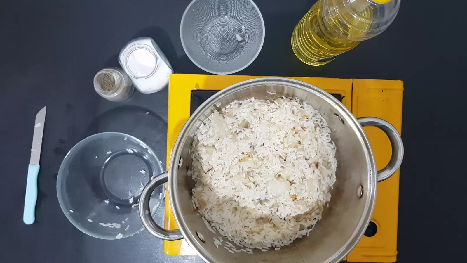 Oil, an empty bowl, a jar of salt, a jar of pepper, a pot with rice, a knife