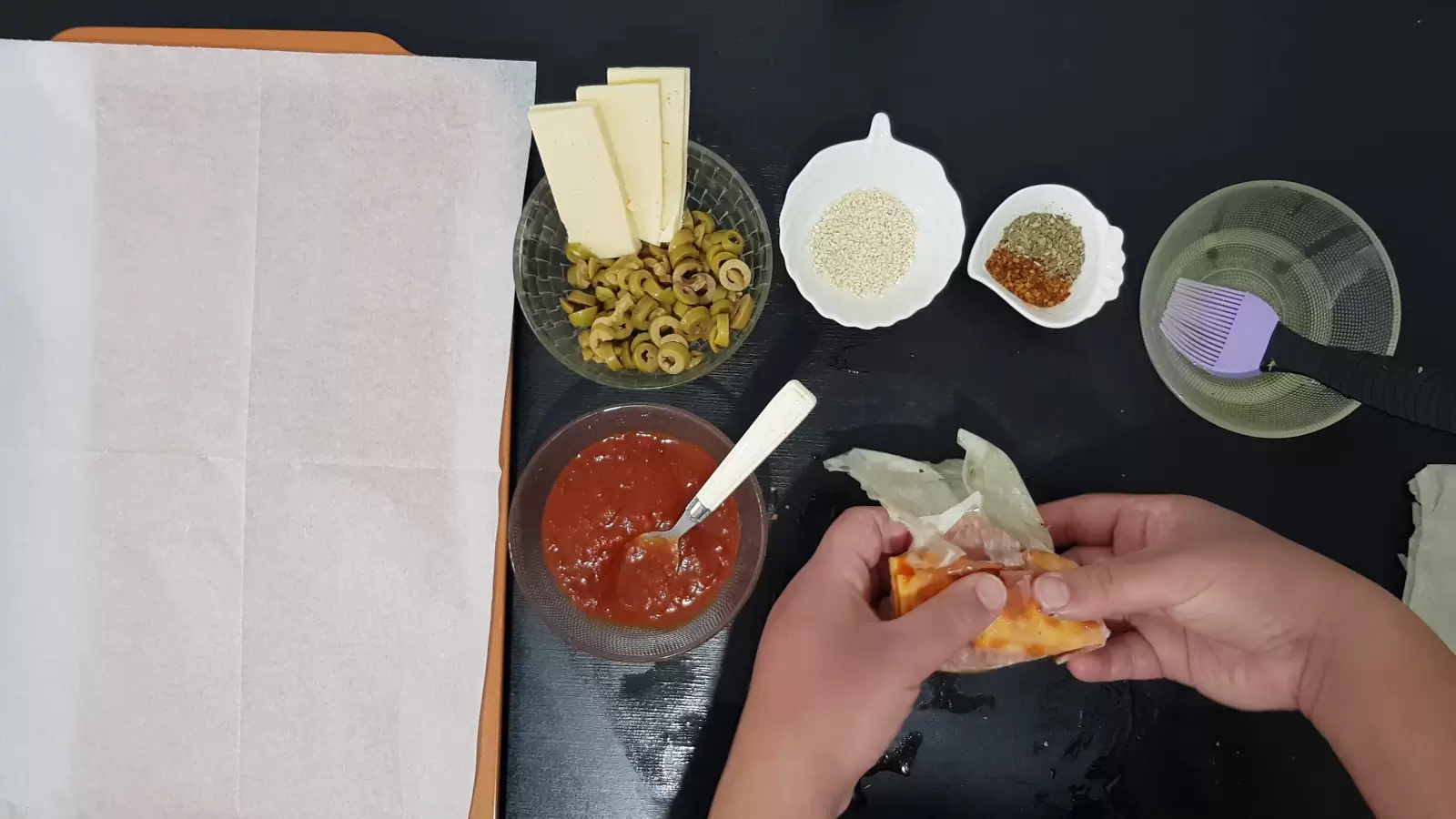 Bowl with oil and brush, bowl with spices, bowl with sesame seeds, bowl with olives and yellow cheese, bowl with tomato sauce, rolled filo dough, spoon and a baking sheet with baking paper