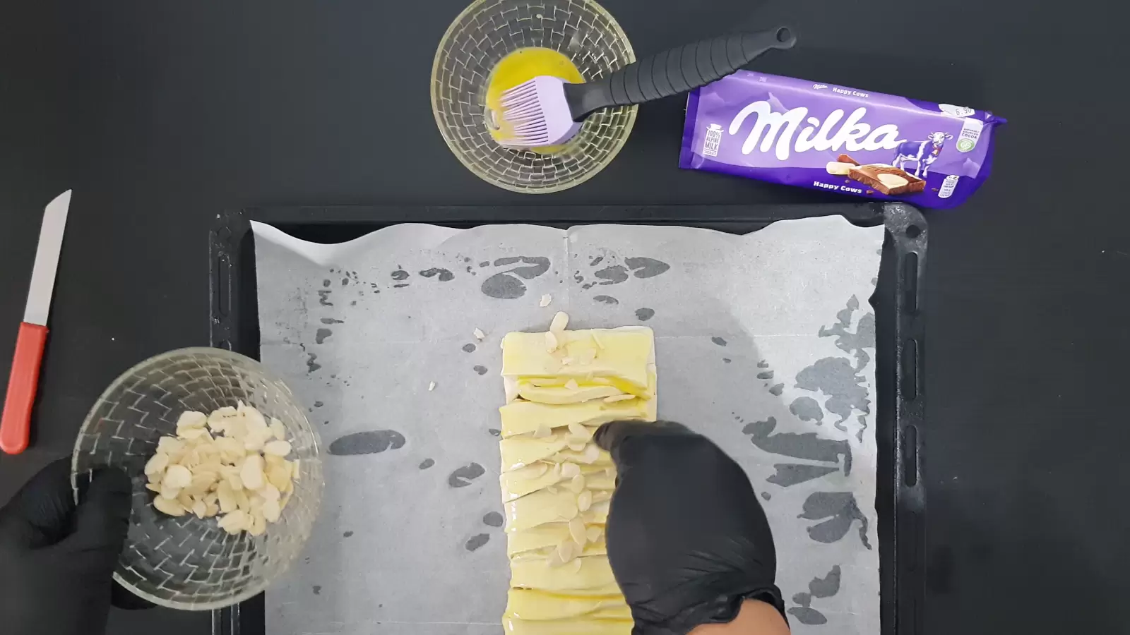 Pack of Milka chocolate, a brush for spreading in a bowl with egg, a bowl with almonds on puff pastry with Milka chocolate on a baking sheet with baking paper, a knife