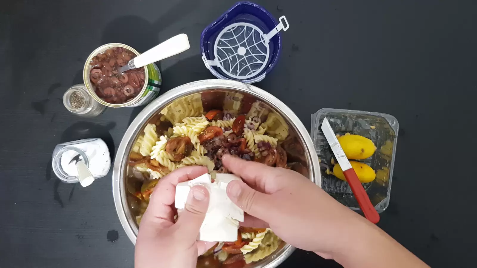 A bowl with pasta and olives, an empty box, Bulgarian cheese, a can with olives and a spoon, a jar of salt, a jar of pepper and a spoon, a box with a lemon and a knife