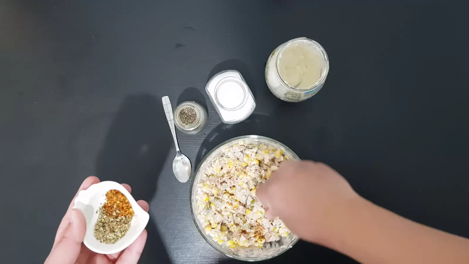 Jar with mayonnaise, jar with salt, jar with black pepper, bowl with chili, spoon, bowl with tuna salad, mayonnaise and corn