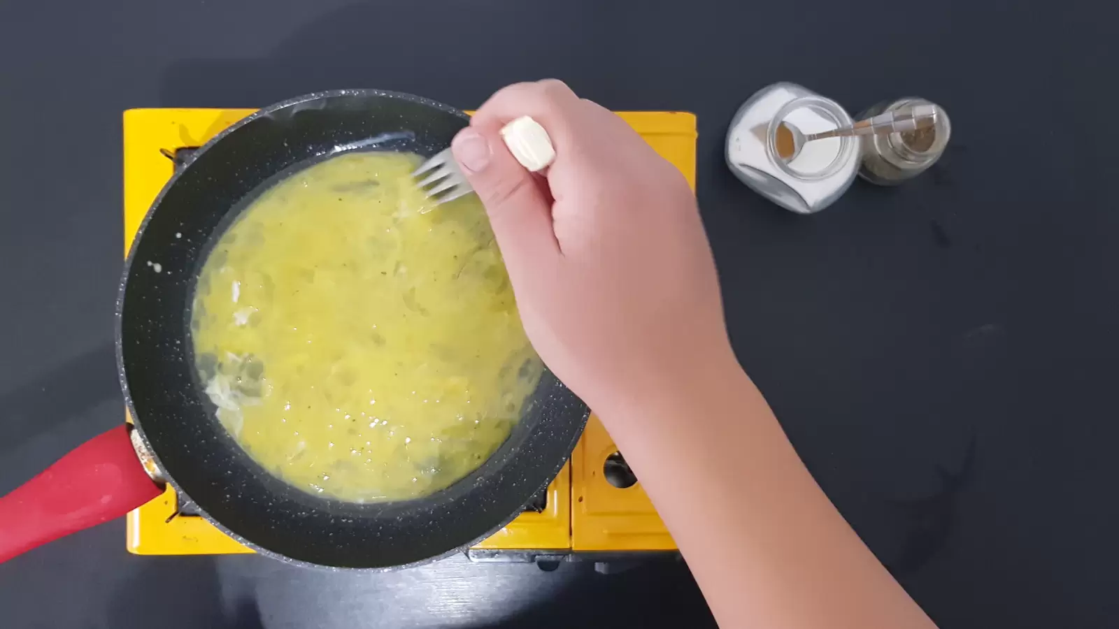 A fork mixes eggs in a pan, a jar of salt with a spoon, a jar of pepper