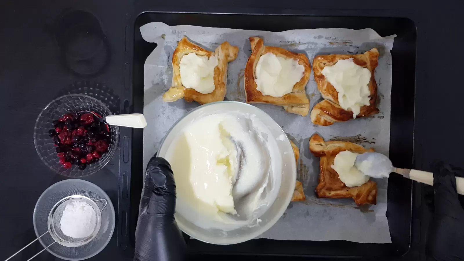 Puff pastry ready on a baking sheet, spoon placing pudding in the center of the pastry, bowl with pudding, bowl with berries, bowl with powdered sugar.