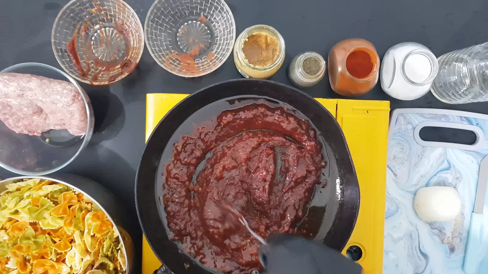 a jar of salt, a jar of red paprika, a jar of black pepper, a jar of crushed garlic, 2 empty bowls, a bowl with ground meat, a bowl with Farfalle pasta, a pan with tomato sauce, a bottle of oil, a cutting board and onions