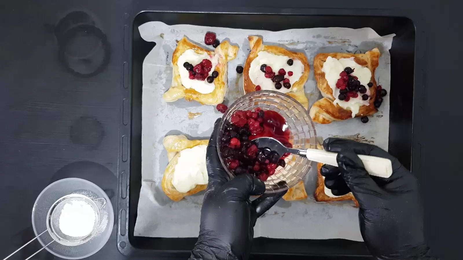 Danish pastry ready on a baking sheet, spoon placing berries in the center of the pastry, bowl with berries, bowl with powdered sugar.