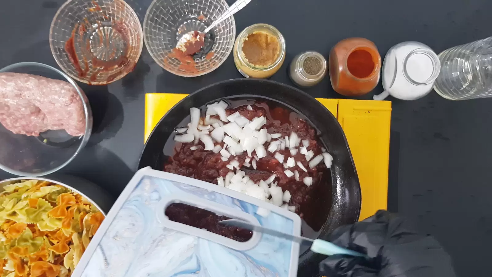 A jar of salt, a jar of red paprika, a jar of black pepper, a jar of crushed garlic, 2 empty bowls, a bowl with ground meat, a bowl with Farfalle pasta, a pan with tomato sauce, a bottle of oil, a cutting board and onions