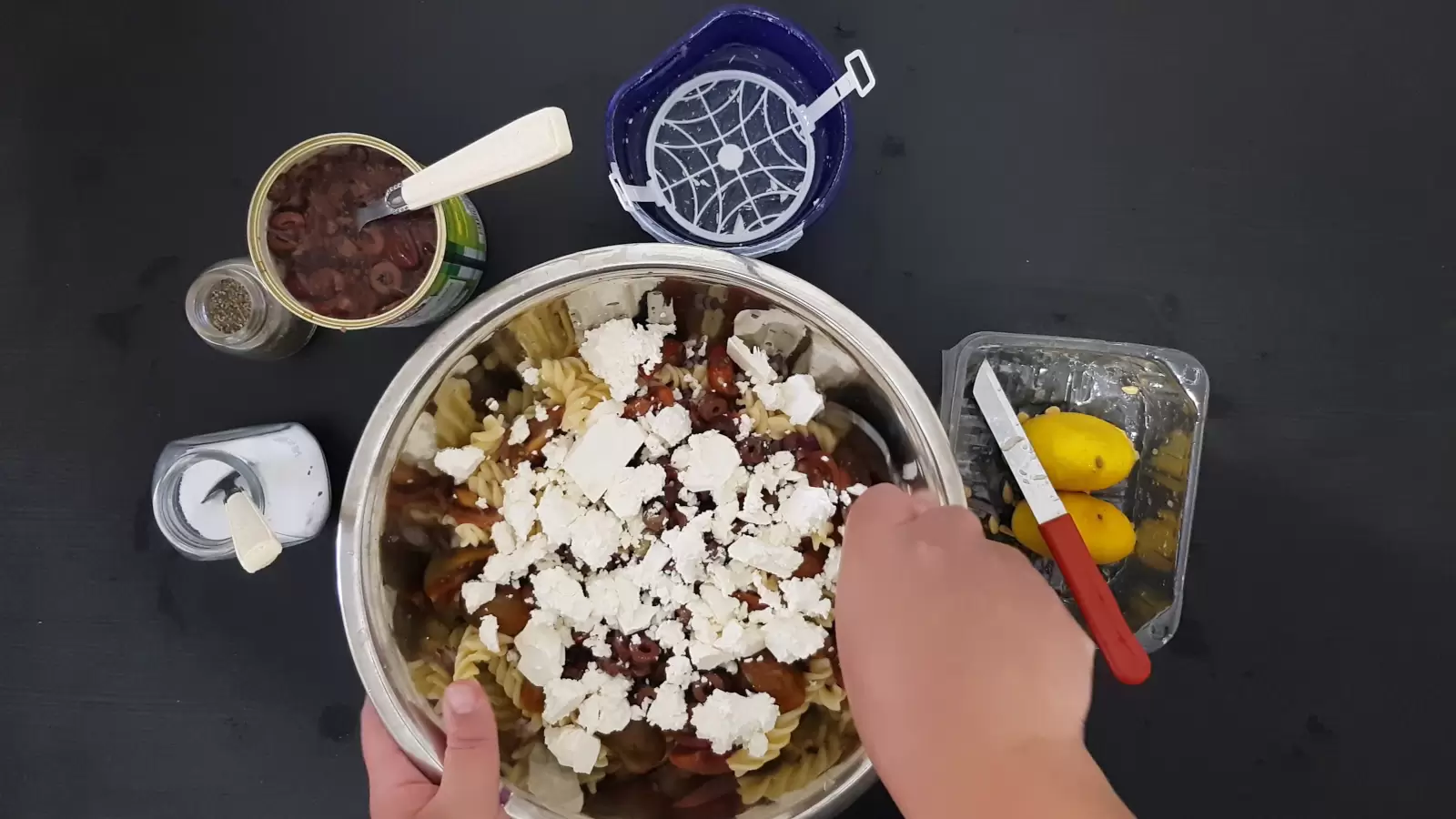 A bowl with Bulgarian cheese pasta and olives, an empty box, a can with olives and a spoon, a jar of salt, a jar of pepper and a spoon, a box with a lemon and a knife