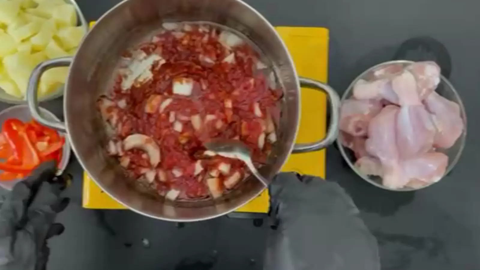 A bowl with chicken thighs, a bowl with potatoes, a bowl with red pepper, a pot with tomato sauce and onions on fire, a spoon