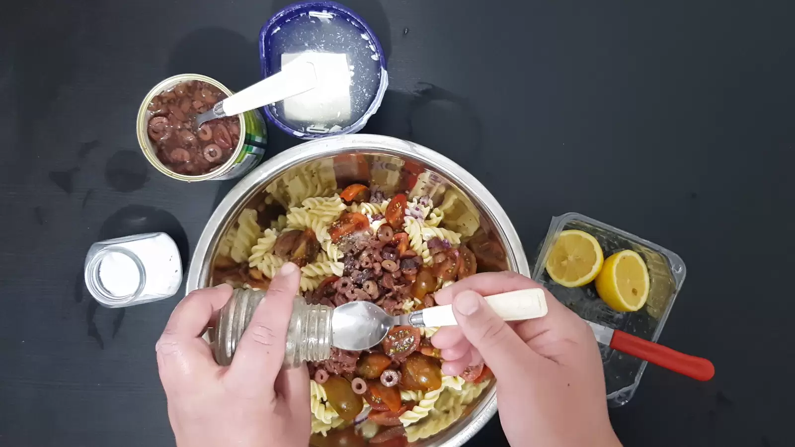 A bowl with pasta and olives, a box with Bulgarian cheese, a can with olives, a jar of salt, a jar of pepper and a spoon, a box with a lemon and a knife