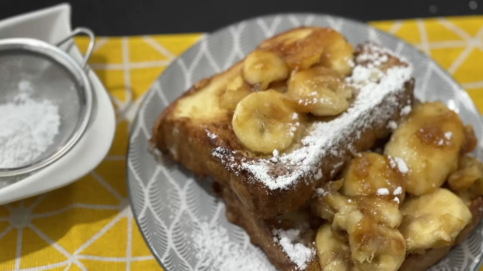 Tasty French toast and caramelized bananas on a plate powdered sugar in a bowl next to it