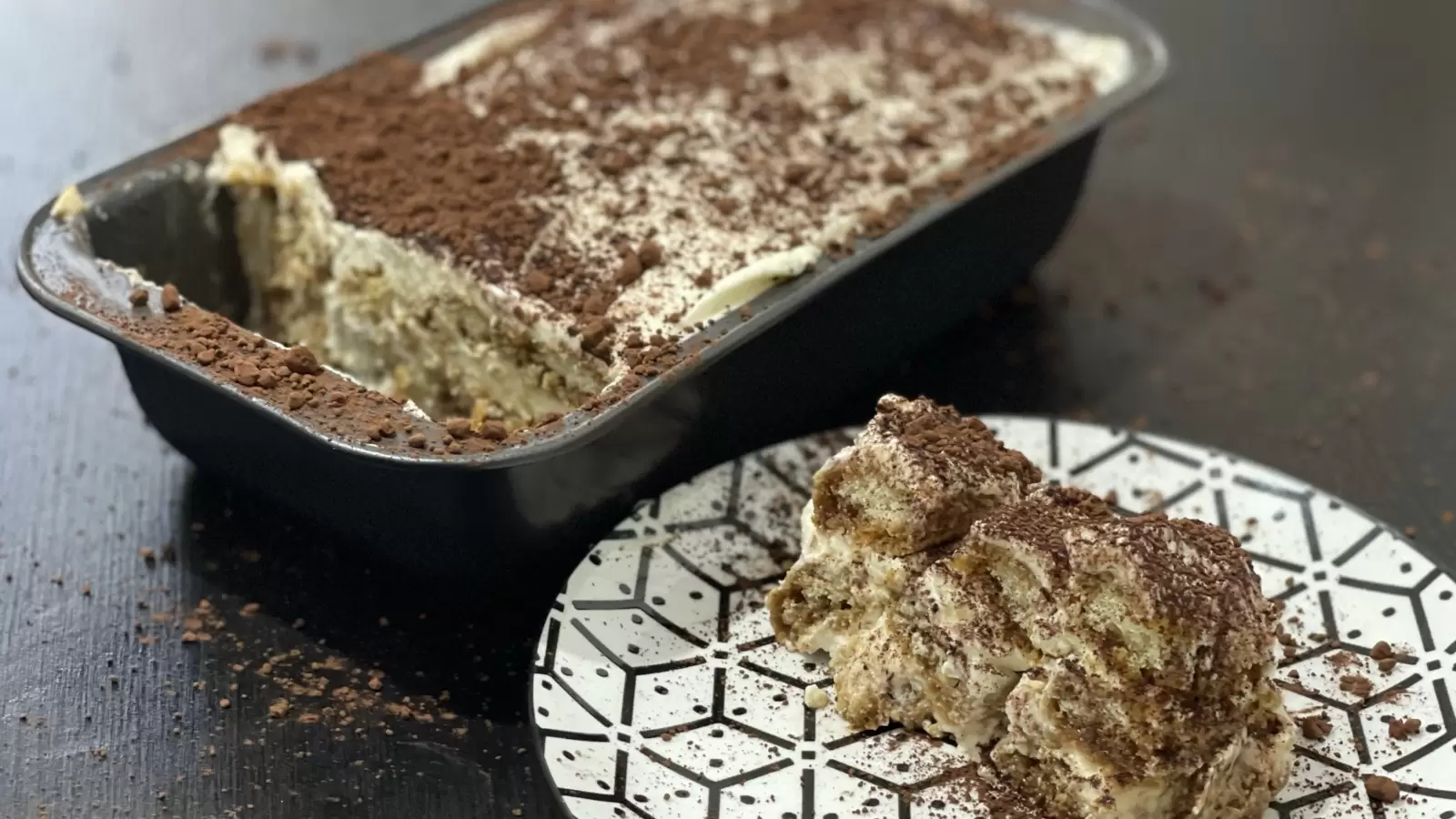 A piece of tiramisu dessert on a plate and a tiramisu cake in a pan next to it