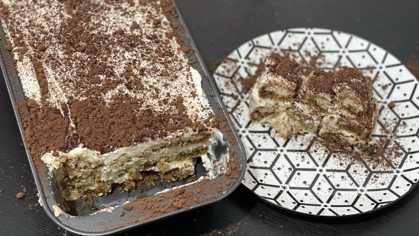 A piece of tiramisu on a plate and a tiramisu cake recipe in a pan next to it.