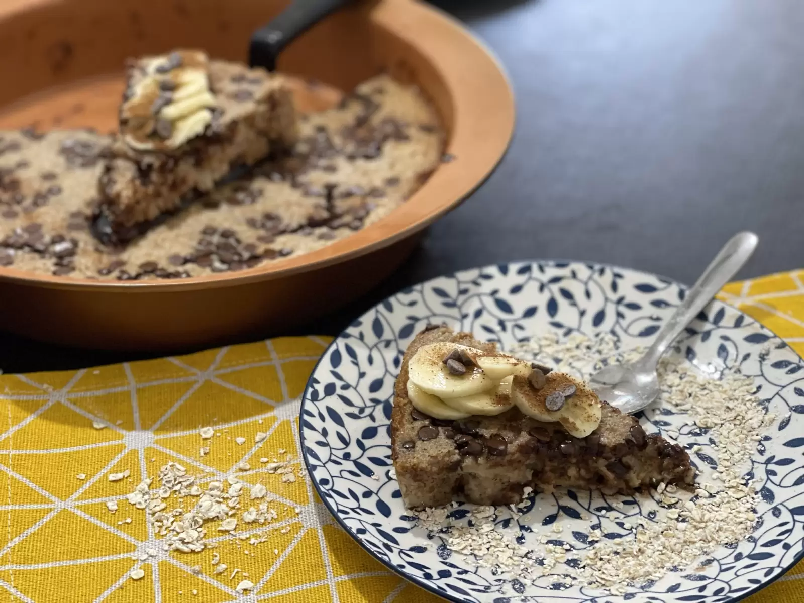 A piece of Oatmeal banana bread on a plate and a spoon with a yellow napkin underneath. Oatmeal scattered. Oatmeal banana bread in a baking dish.